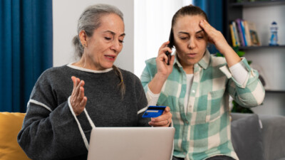 Two distraught women, one of whom is on the phone while the other holds a credit card and laptop.