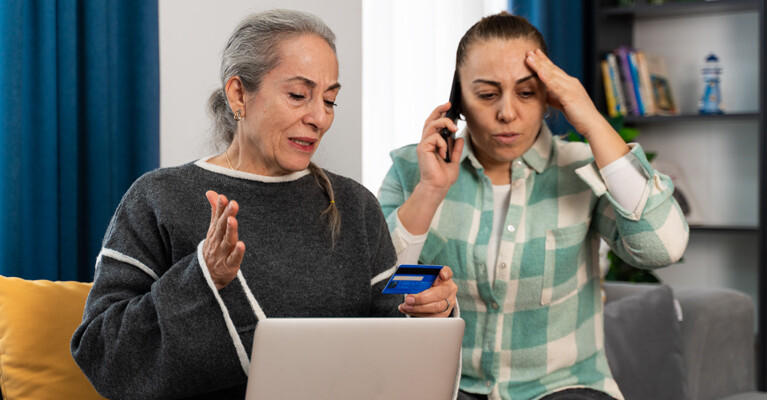 Two distraught women, one of whom is on the phone while the other holds a credit card and laptop.