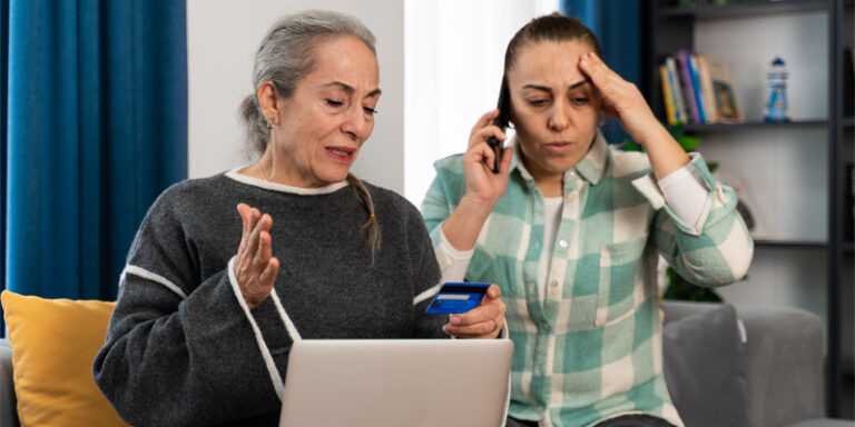 Two distraught women, one of whom is on the phone while the other holds a credit card and laptop.