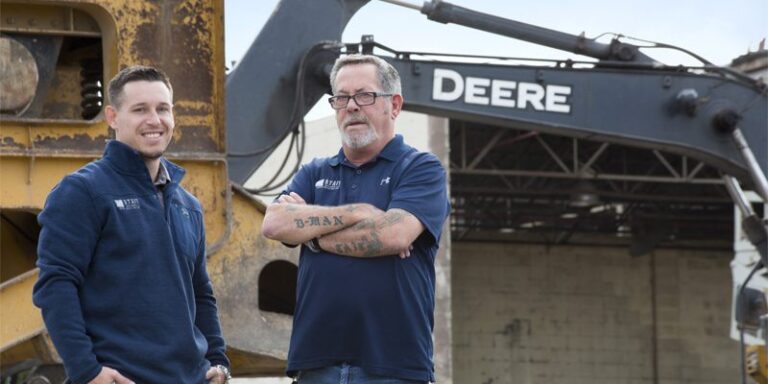 Two men in front of construction equipment.