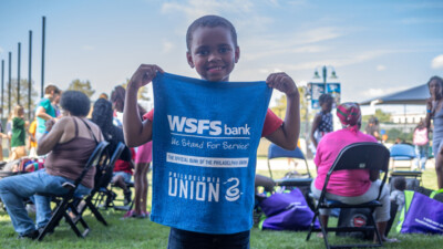 A child at the Philadelphia Union Backpack Carnival holding a rally towel that says “WSFS Bank. We Stand for Service. The Official Bank of the Philadelphia Union.”