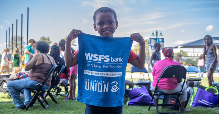 A child at the Philadelphia Union Backpack Carnival holding a rally towel that says “WSFS Bank. We Stand for Service. The Official Bank of the Philadelphia Union.”
