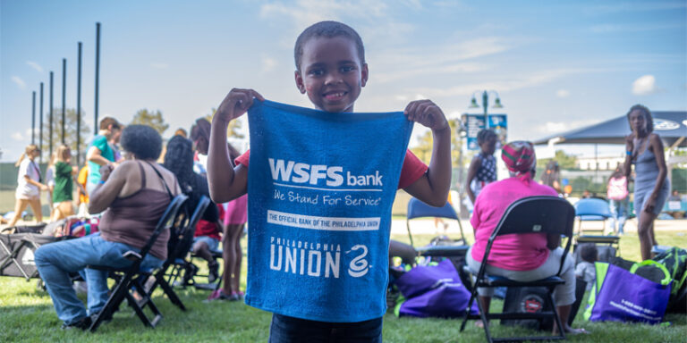 A child at the Philadelphia Union Backpack Carnival holding a rally towel that says “WSFS Bank. We Stand for Service. The Official Bank of the Philadelphia Union.”