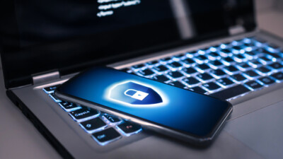 A cell phone with a blue lock and shield icon, laying on top of a laptop computer.