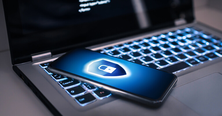 A cell phone with a blue lock and shield icon, laying on top of a laptop computer.