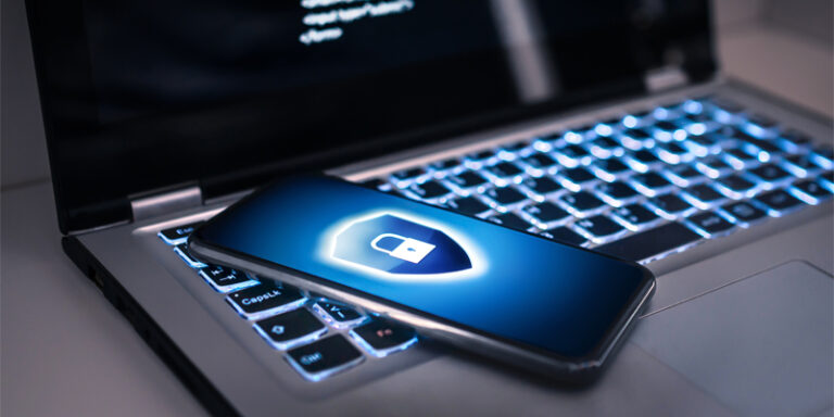 A cell phone with a blue lock and shield icon, laying on top of a laptop computer.