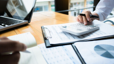 Hands holding pens, on top of a desk covered in paperwork and charts.