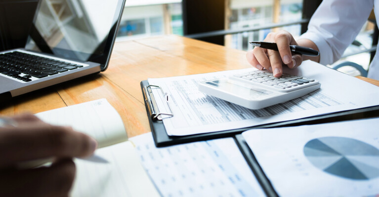 Hands holding pens, on top of a desk covered in paperwork and charts.