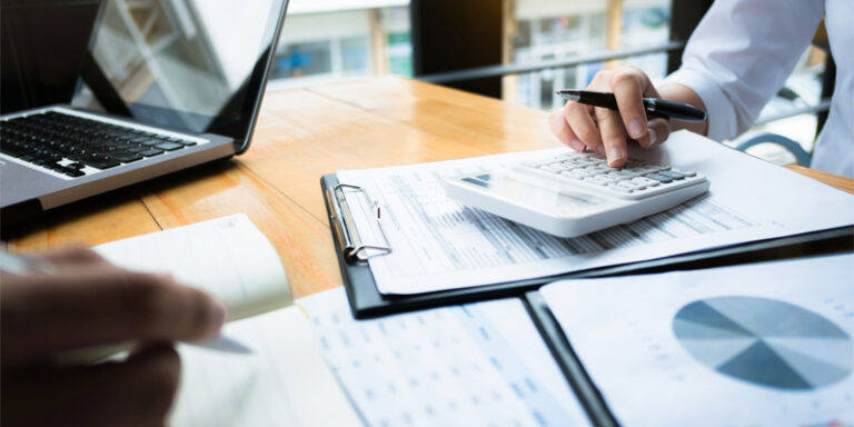 Hands holding pens, on top of a desk covered in paperwork and charts.