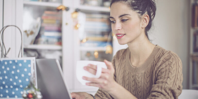 A woman drinking from a mug and using a laptop.