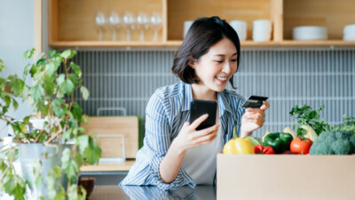 A woman holding her cell phone and her credit card, surrounded by vegetables and houseplants.