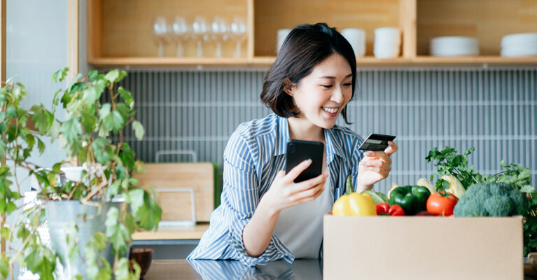 A woman holding her cell phone and her credit card, surrounded by vegetables and houseplants.