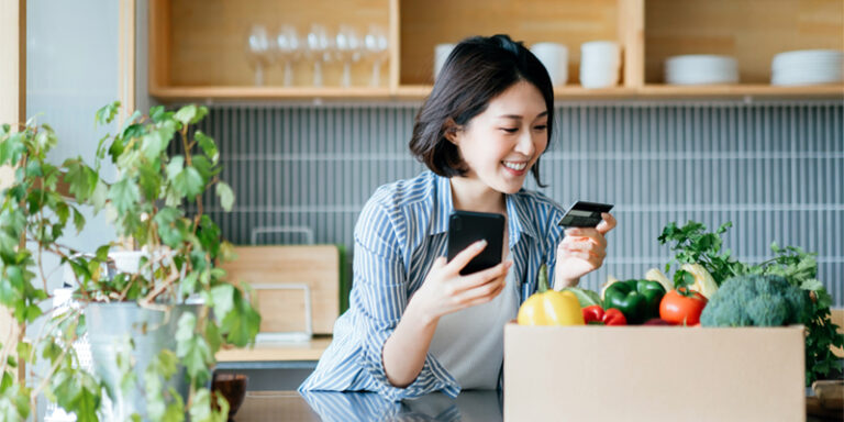 A woman holding her cell phone and her credit card, surrounded by vegetables and houseplants.