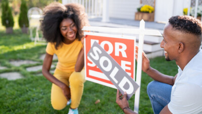 A man and a woman happily putting a "SOLD" banner over a For Sale sign, in the front yard of their recently purchased house.