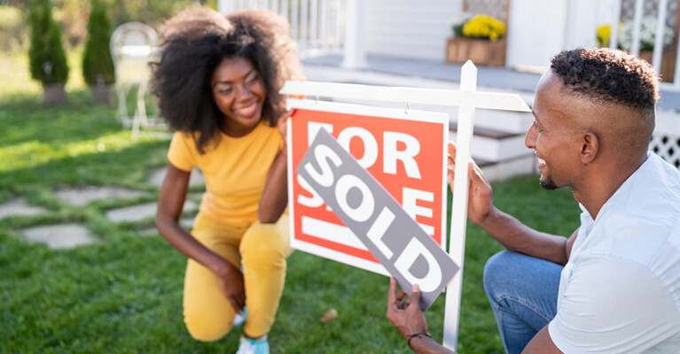 A man and a woman happily putting a "SOLD" banner over a For Sale sign, in the front yard of their recently purchased house.