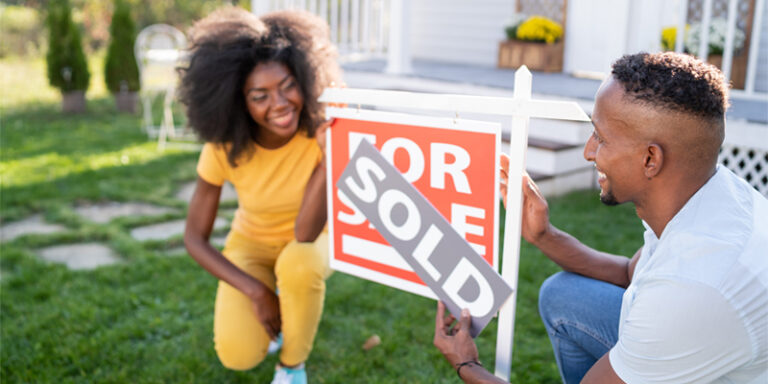 A man and a woman happily putting a "SOLD" banner over a For Sale sign, in the front yard of their recently purchased house.