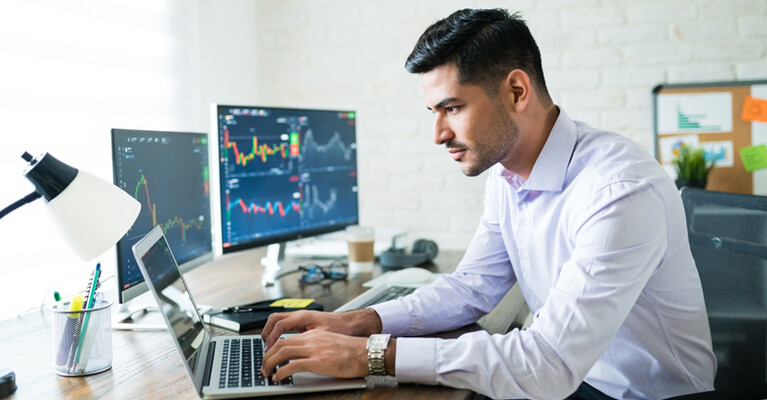 Man using laptop computer, with stock charts on monitors.