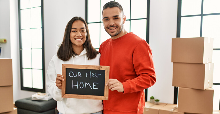 Two people holding a chalkboard that reads "Our First Home."