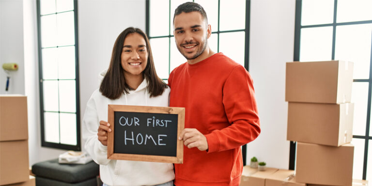 Two people holding a chalkboard that reads "Our First Home."