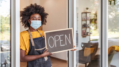 A business owner holding an "Open" sign in the front doorway of their business.