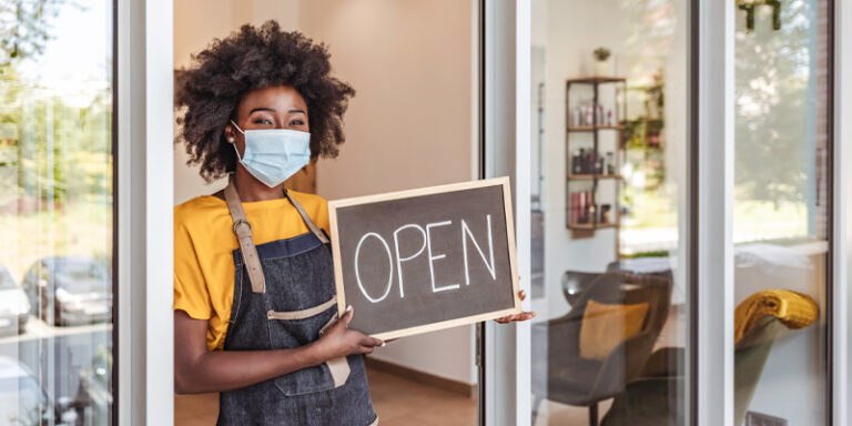 A business owner holding an "Open" sign in the front doorway of their business.