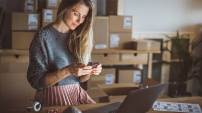 A woman using her laptop and cell phone, packaged orders behind her.