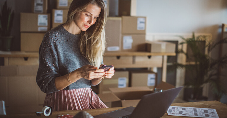 A woman using her laptop and cell phone, packaged orders behind her.