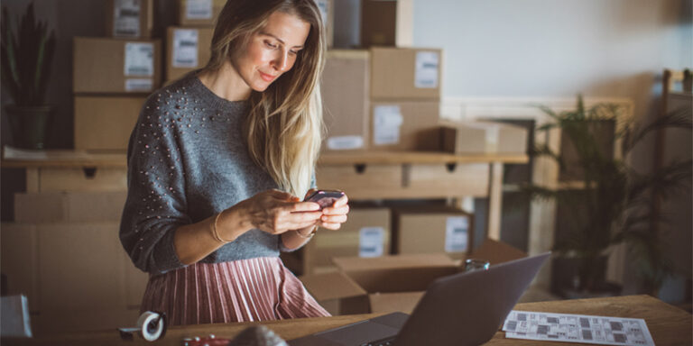 A woman using her laptop and cell phone, packaged orders behind her.