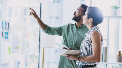 A man and a woman, brainstorming, at a board covered in sticky notes and papers.