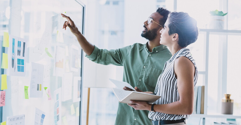 A man and a woman, brainstorming, at a board covered in sticky notes and papers.