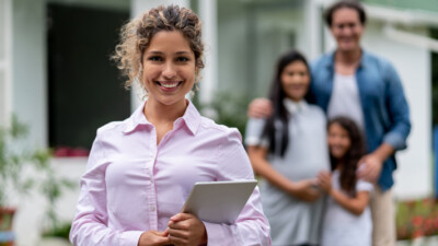 Real estate agent holding tablet, smiling with a family in front of a house.