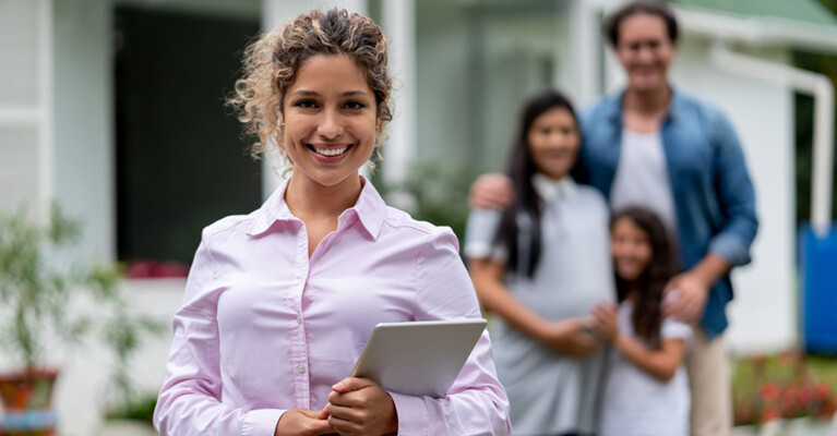 Real estate agent holding tablet, smiling with a family in front of a house.