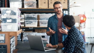 A man and a woman, talking, while surrounded by products in a warehouse.