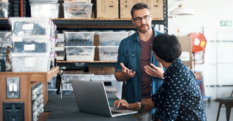 A man and a woman, talking, while surrounded by products in a warehouse.