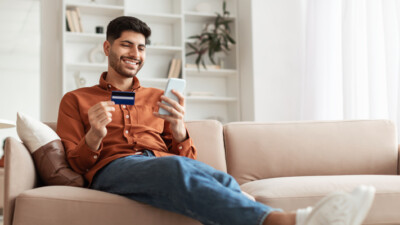 Man relaxing on the couch looking at bank card and cell phone.