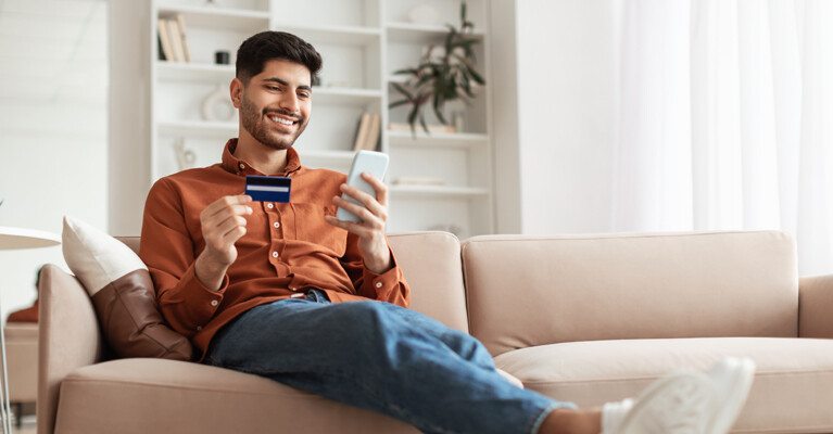 Man relaxing on the couch looking at bank card and cell phone.