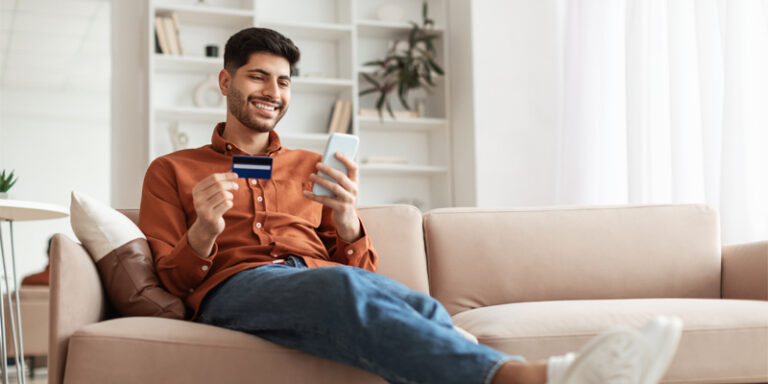 Man relaxing on the couch looking at bank card and cell phone.