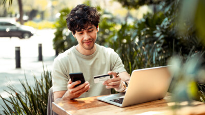 A man looking at his cell phone and his credit card, while using his laptop.