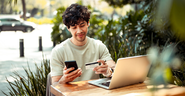 A man looking at his cell phone and his credit card, while using his laptop.