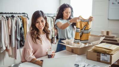 Two women packaging clothing orders and using a laptop.