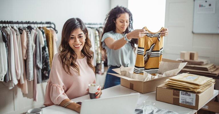 Two women packaging clothing orders and using a laptop.