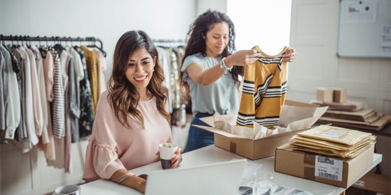 Two women packaging clothing orders and using a laptop.