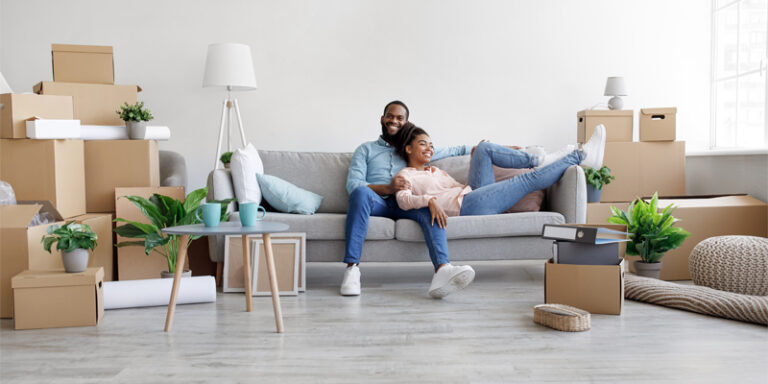 A couple relaxing on the couch in their new home, amidst unpacked boxes.