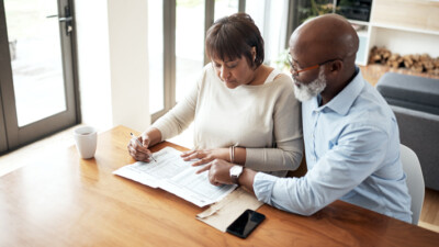 A man and a woman filling out paperwork together at their kitchen table.