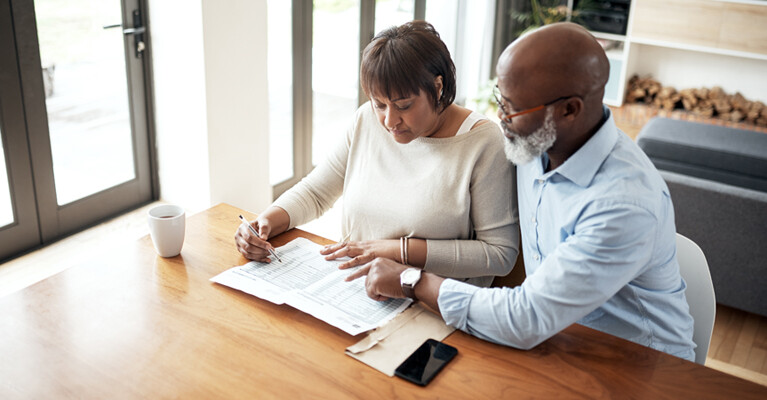 A man and a woman filling out paperwork together at their kitchen table.