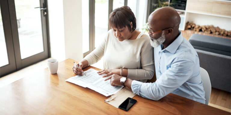 A man and a woman filling out paperwork together at their kitchen table.