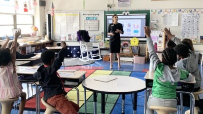 An early education classroom with several children raising their hands.