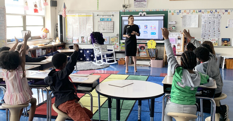 An early education classroom with several children raising their hands.