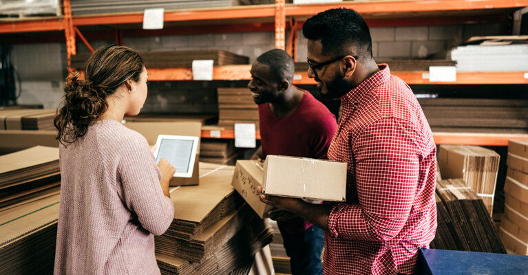 Woman and two men in a warehouse, looking at a tablet and holding cardboard boxes.
