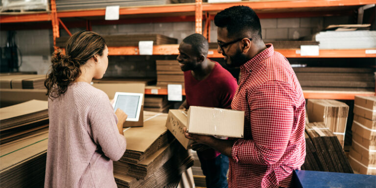 Woman and two men in a warehouse, looking at a tablet and holding cardboard boxes.